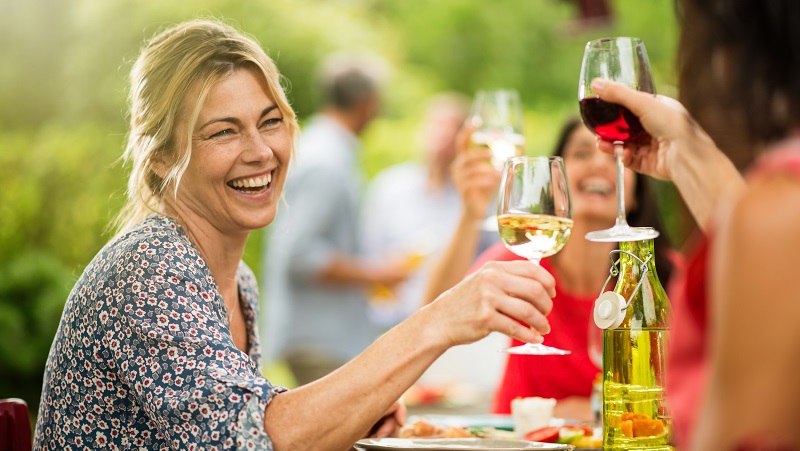 Leende kvinna skålar med vin i solig trädgårdsmiljö omgiven av vänner och grönskande bakgrund.